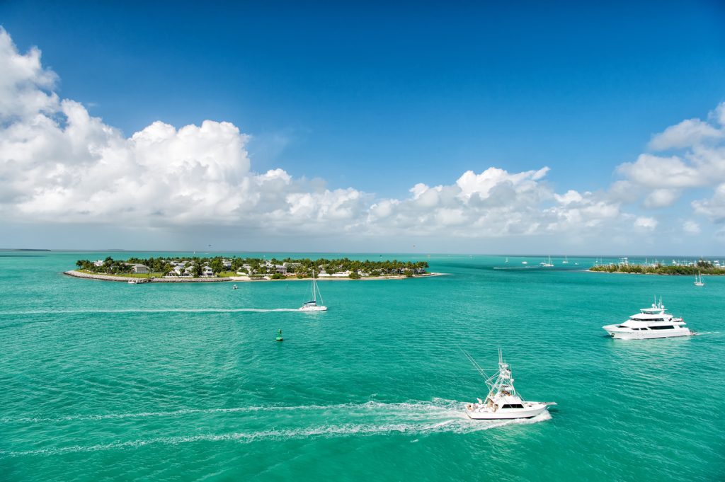 Cruise touristic boats or yachts floating by island with houses and green trees on turquoise water and blue cloudy sky, yachting and isle life around beautiful Key West Florida, USA. FloridaKeysVillas.com